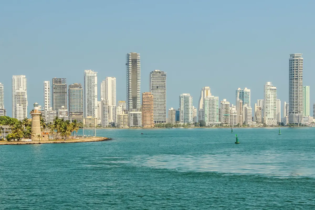 Bílý maják na zeleném výběžku, za nímž se přes tyrkysovou vodu tyčí vysoké věžové domy čtvrti Bocagrande.