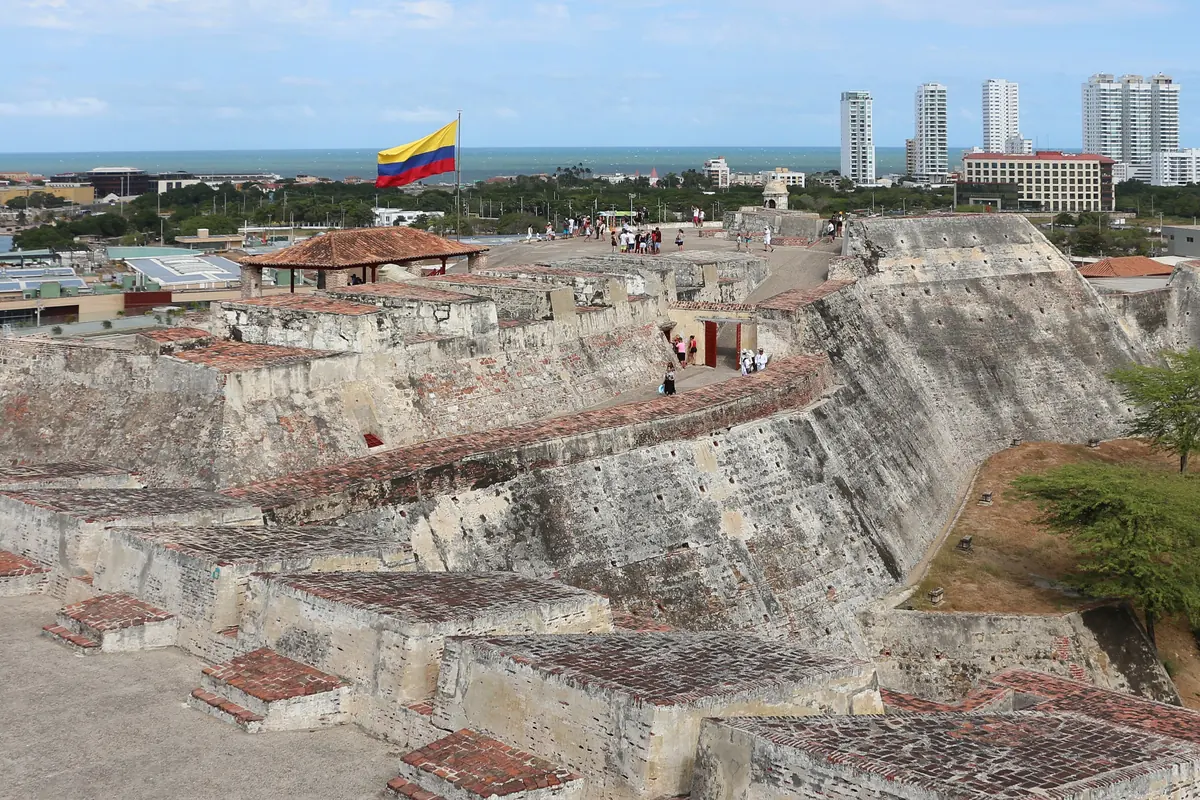 Šedé kamenné hradby Castillo San Felipe de Barajas s kolumbijskou vlajkou a siluetou Cartageny v pozadí.