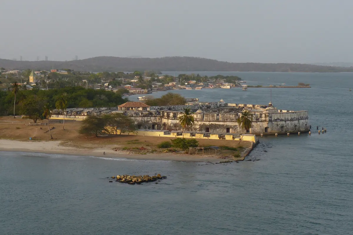 Et kystfort med lave mure ved en strand og roligt vand under en grå himmel